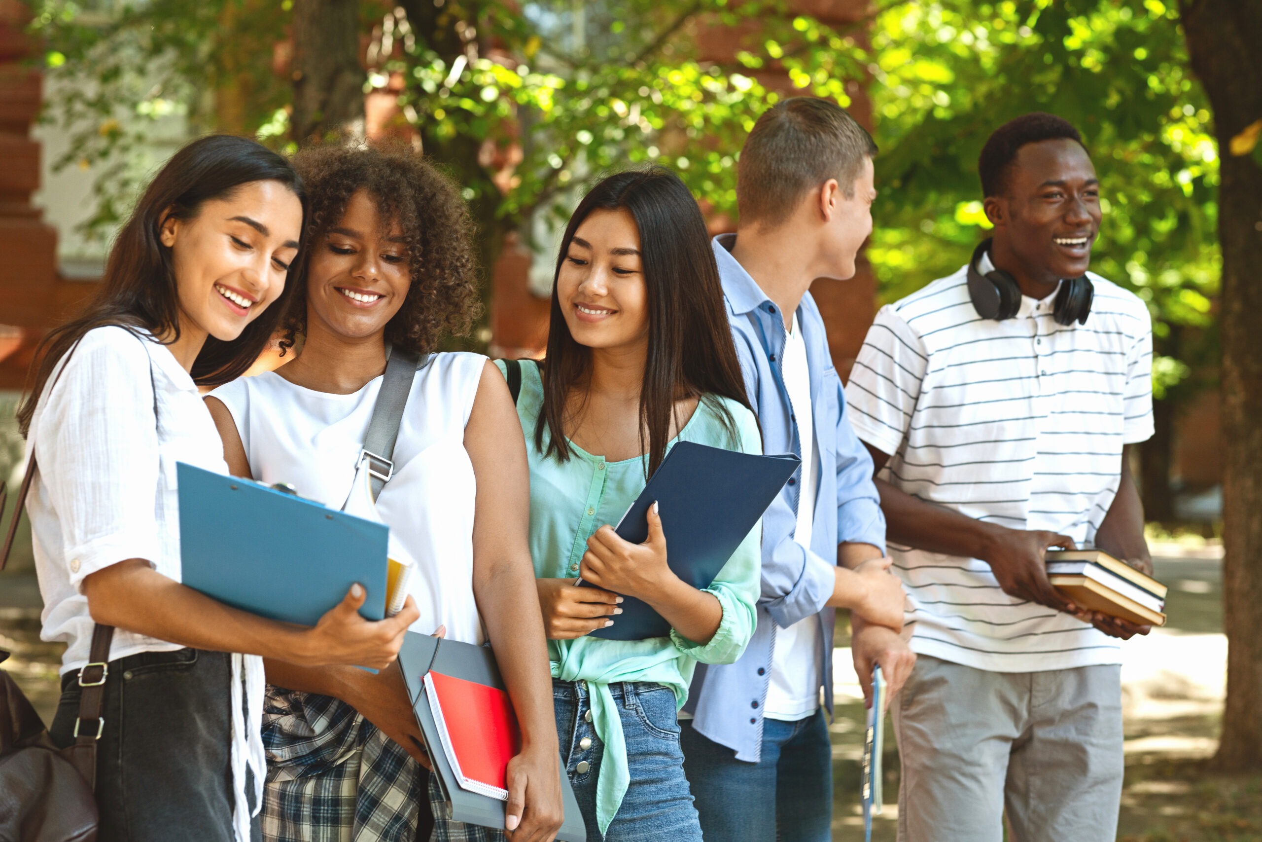 Three smiling students share notes and laughter outdoors, while two peers engage in conversation nearby, highlighting a vibrant campus atmosphere.